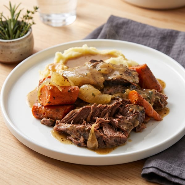 Steaming pot roast with carrots, onions, and gravy, served with mashed potatoes on a white plate.