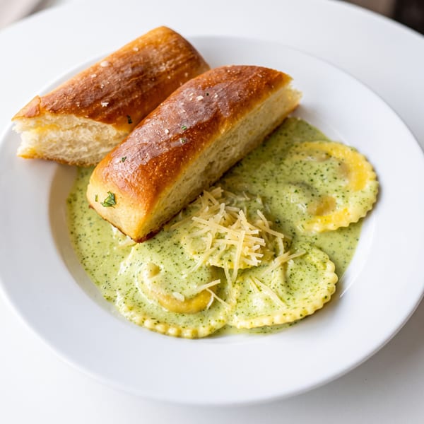 Ravioli in a creamy green sauce, topped with shredded cheese and two breadsticks on a white plate.