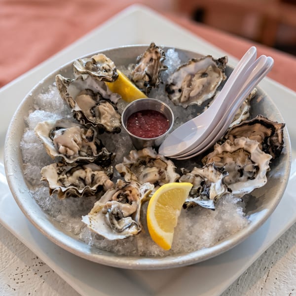 A dozen fresh oysters on a bed of ice with lemon wedges, cocktail sauce, and spoons.